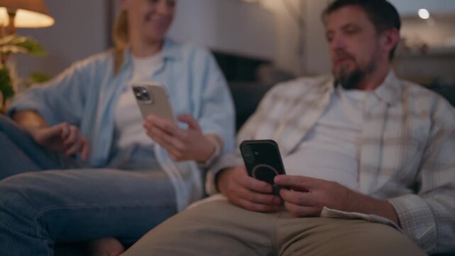 Couple sitting side by side on a sofa and speaking without looking up from their phones, representing cold communication, emotional distance, relationship difficulties and lack of connection.