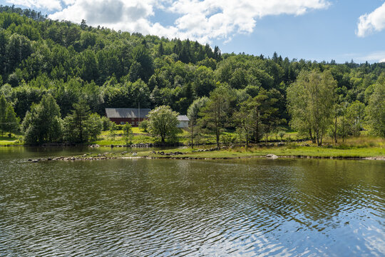 View of a traditional red wooden house nestled in a lush green forest along the calm waters of a lake in Mandal, Agder, Norway.