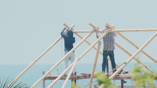 Two builders constructing a wooden roof structure with the blue sea in the background