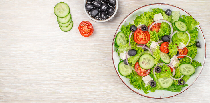 Fresh vegetable salad with cucumbers, tomatoes, olives, and feta cheese served on a white plate, with sliced cucumbers and a bowl of olives on a wooden table