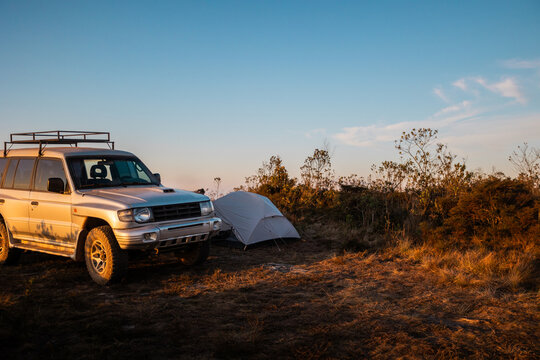 Acampamento de aventura ao p&ocirc;r do sol em um cume de montanha com vista panor&acirc;mica, ve&iacute;culo 4x4, barracas e cadeira de acampamento em uma paisagem natural acidentada.