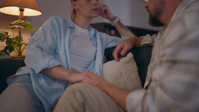 Couple sitting on a sofa and having a tense conversation at home, showing emotional distance, relationship difficulties, communication problems and unresolved conflict.