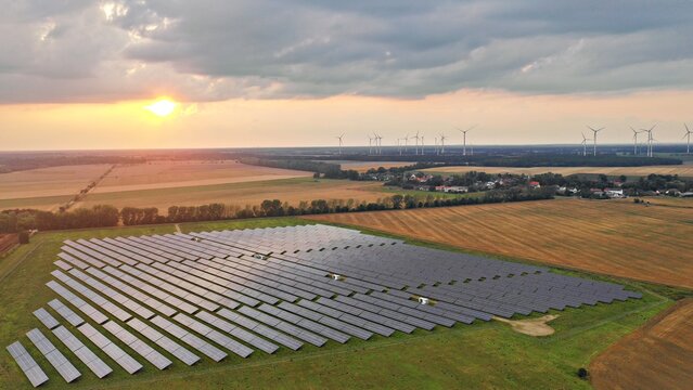 Aerial view of a solar farm with rows of photovoltaic panels set in a rural landscape with wind turbines in the distance under a sunset sky in Markisch-Oderland, Brandenburg, Germany.