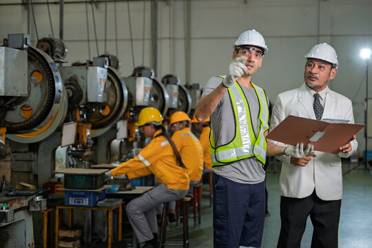 Senior Asian factory manager in a suit reviewing production documents with a male industrial engineer wearing safety gear on the manufacturing floor with other workers in the background.
