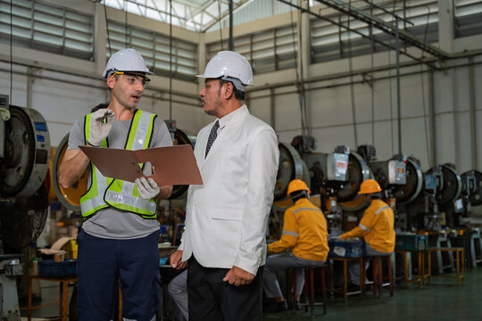 Senior Asian factory manager in a suit reviewing production documents with a male industrial engineer wearing safety gear on the manufacturing floor with other workers in the background.