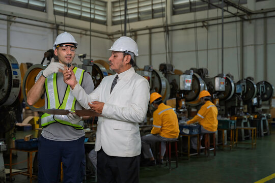 Senior Asian factory manager in a suit reviewing production documents with a male industrial engineer wearing safety gear on the manufacturing floor with other workers in the background.