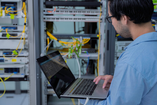 Focused Asian male IT network administrator uses a laptop to monitor server performance inside a bright data center managing complex corporate cybersecurity operations and infrastructure.