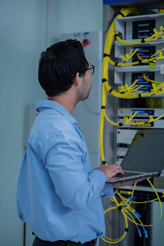 Focused Asian male IT network administrator uses a laptop to monitor server performance inside a bright data center managing complex corporate cybersecurity operations and infrastructure.
