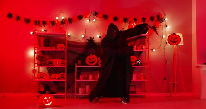 Funny person wearing grim reaper costume dancing in spooky Halloween decorated room with red lighting. Mysterious figure in black hooded cloak holding scythe, having fun among glowing jack-o-lanterns.