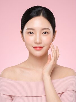 Beautiful young asian woman with a pink facial clay mask on her skin looking at the camera with a soft smile and touching her cheek