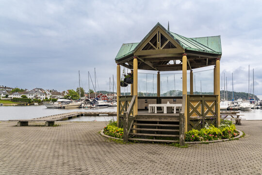 View of the wooden gazebo in the harbor surrounded by flowers with boats docked in the background in Lillesand, Agder, Norway.