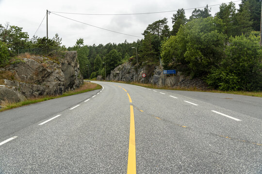 View of a winding asphalt road flanked by rocky cliffs and dense forest under an overcast sky in Lillesand, Agder, Norway.