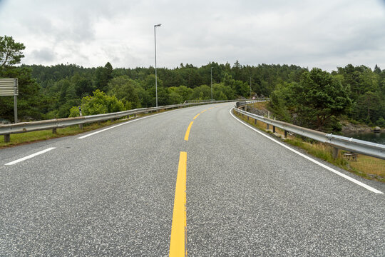 View of a winding asphalt road with yellow lane markings, flanked by metal guardrails and lush green forest under a cloudy sky in Lillesand, Agder, Norway.