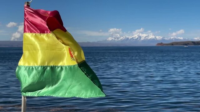 Bandeira da Bol&iacute;via a ondear ao vento com o Lago Titicaca e a Cordilheira Real ao fundo. Vista c&eacute;nica das montanhas nevadas dos Andes sob c&eacute;u azul.