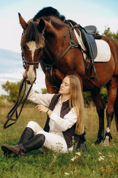 Young beautiful female jockey sits on a meadow near her horse at sunset.