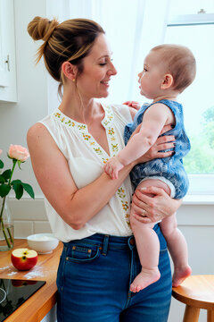 Vertical shot of Caucasian young adult mother holding baby, smiling and bonding in kitchen, showing caregiving and family life. Ideal for parenting, childcare, maternity, home routine marketing