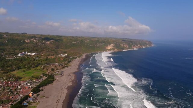 Aerial view of Pantai Parangkusumo featuring dark sand beach, rolling ocean waves, and lush cliffs in Kecamatan Kretek, Daerah Istimewa Yogya, Indonesia.