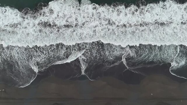 Ocean waves roll onto a black volcanic sand beach, creating white foam patterns along the tropical Pacific coastline.