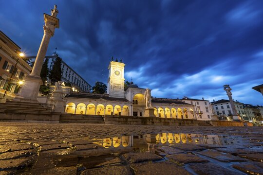 Illuminated main square Piazza della Libert&agrave; with Loggia e Tempietto di San Giovanni and clock tower, Castello di Udine behind, blue hour, reflection in water puddle, dramatic sky, old town, Udine, Friuli-Venezia Giulia, Italy