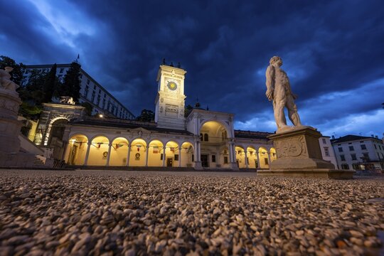 Illuminated main square Piazza della Libert&agrave; with Loggia e Tempietto di San Giovanni and clock tower, Castello di Udine in the back, statue of Ercole e Caco, blue hour, dramatic sky, old town, Udine, Friuli-Venezia Giulia, Italy