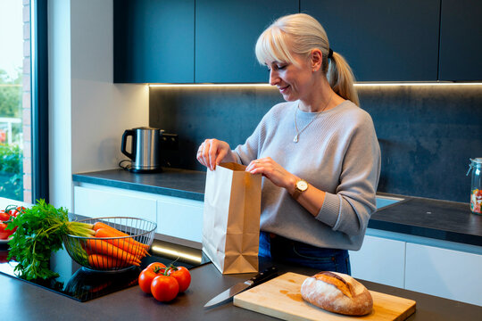 Middle aged Caucasian woman unpacking grocery paper bag in kitchen beside fresh vegetables and bread, showing healthy meal planning. Ideal for nutrition, home cooking, food retail content