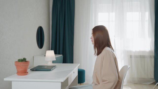 Engaged white woman at study desk, sits and opens phone and laptop while browsing grammar exercises, typing notes and joining virtual tutor call, focused expression, bright curtains and desk lamp,
