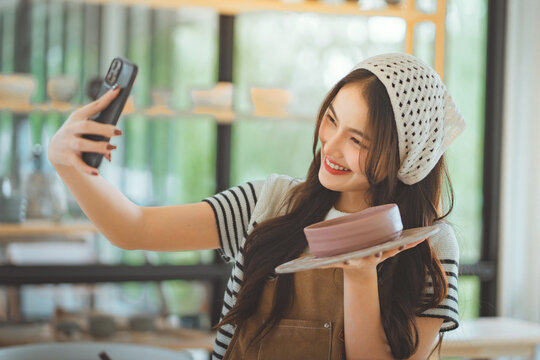 Happy asian female artist taking selfie with handmade pottery design in creative studio &ndash; smiling sculptor sharing artwork on social media for online selling.