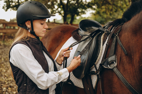 A young woman jockey adjusts the harness on her horse before training.