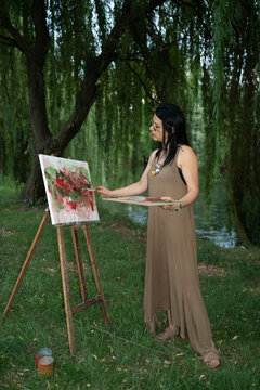 Elegant woman artist painter by the river painting on a canvas on an easel outdoors and holding paintbrushes and an artist palette