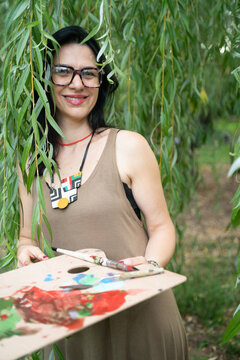 Young woman artist with artist palette by the willow tree in the forest, looking at the camera and smiling