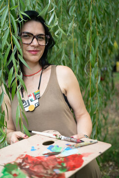 Young woman artist with artist palette by the willow tree in the forest, looking at the camera and smiling