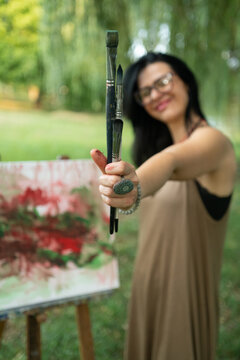 Portrait of a happy woman artist by the canvas outdoors, holding paintbrushes and smiling, close-up on brushes