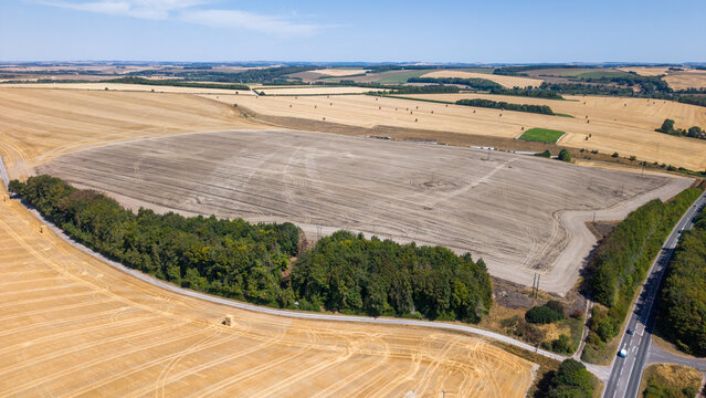 Aerial view of harvested fields and arable farmland in the Wiltshire countryside with dry stubble, hay bales, and a road winding through the landscape in Salisbury, England, United Kingdom.