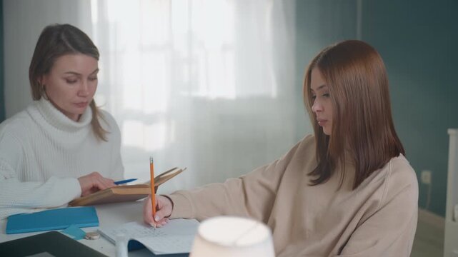 White coach guiding asian student, tutor in white sweater reviewing notes and giving feedback at home desk, pencil and notebook exchange, warm lamp glow and small plant on table, constructive