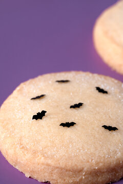 Close-up photo of a Halloween sugar cookie with black bat sprinkles on a purple surface. Shot at a 45 degree angle with soft selective focus