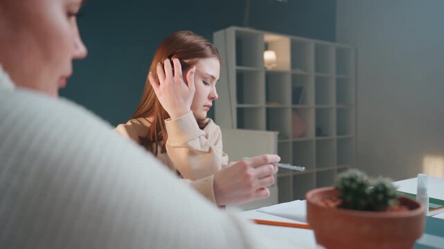 Tutor guiding student at study table during focused practice, student writing on worksheet while tutor observes, soft lamp glow and potted plant on table, bookshelf backdrop, steady coaching energy