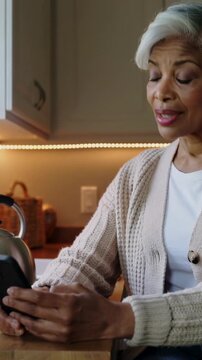 Vertical video: Holding smartphone senior woman in cardigan tapping at kitchen counter with kettle