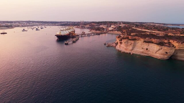 Drone flies forward over Marsaxlokk Bay at sunset, passing the illuminated LNG terminal toward the historic Delimara Battery perched on limestone cliffs.