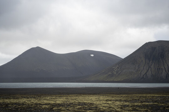 View of volcanic mountains rising above a calm lake with mossy foreground under an overcast sky in Iceland.