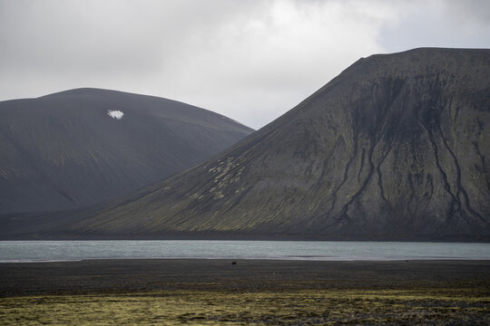 View of volcanic mountains with dark slopes and patches of green moss rising above a calm lake in Iceland.