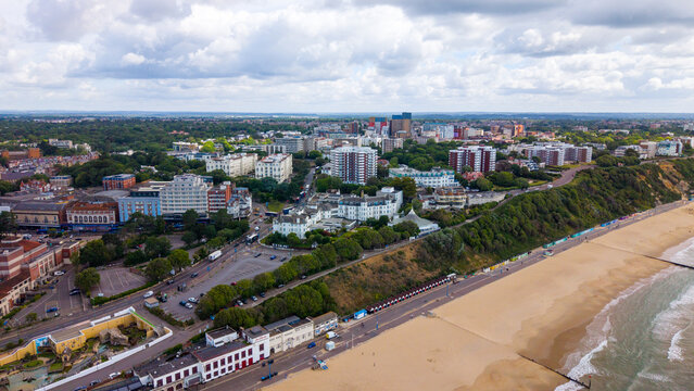 Aerial view of Bournemouth Pier and the sandy beach stretching along the coastline with urban buildings, lush trees, and a cloudy sky in Bournemouth, England, United Kingdom.