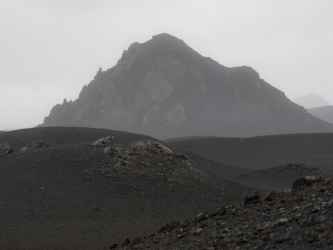 View of a rugged volcanic landscape with dark sand dunes and a misty mountain peak under an overcast sky in Iceland.