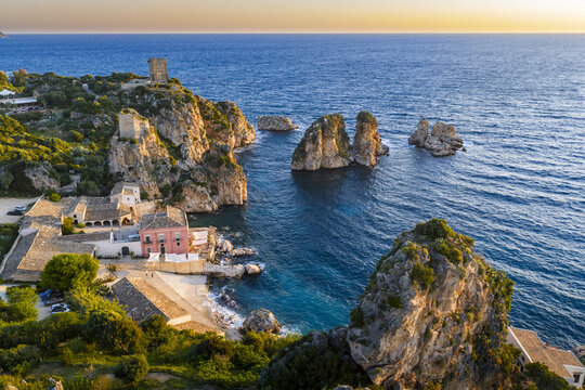 Aerial view of Tonnara di Scopello with its historic buildings, rocky cliffs, and sea stacks rising from the clear blue Mediterranean water at sunset in Scopello, Sicily, Italy.