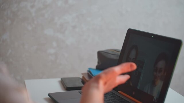 Two girls tutoring online at laptop, focused home desk scene with grammar coach on video call, student practices pronunciation and sentence structure, notebooks and textbooks stacked beside laptop,