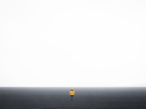 View of a person wearing a yellow jacket standing alone in a vast, dark volcanic landscape under a bright, overcast sky in Iceland.