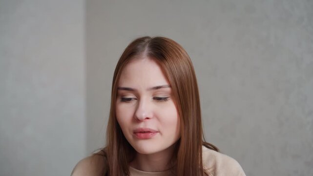 Caucasian young woman on webcam speaking clearly, practicing pronunciation and grammar for online presentation, closeup facial expressions, laptop camera framing, neutral home wall background, focused