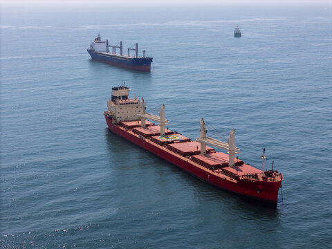 Aerial view of two large cargo ships sailing in the open ocean, with a red bulk carrier in the foreground and a dark blue vessel in the distance near Cascais, Lisboa, Portugal.