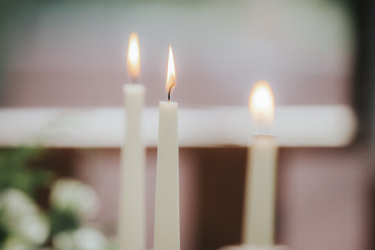 Three lit white candles with soft flames on a blurred background, surrounded by greenery and floral elements, creating a serene and peaceful atmosphere