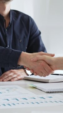 Business partners shaking hands after successful meeting in office while another person is clapping hands over financial documents, laptop and notebook