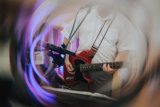 Male guitarist wearing a white shirt plays electric guitar with microphone in front, blurred background with colorful lights and musical equipment visible